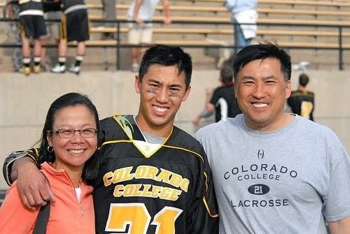Three people smiling and standing together outdoors; one is a woman wearing glasses and a bright orange hoodie, the other two are men, one in a Colorado College lacrosse shirt and the other in a black and yellow sports jersey, with a stadium or bleac
