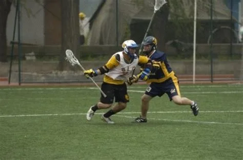 Two male lacrosse players in action on a field, one in a yellow and black uniform and helmet, the other in a blue and yellow uniform and helmet, competing for the ball.