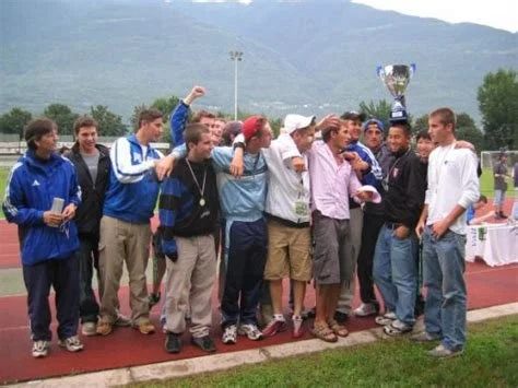 Group of young men standing on a track field, celebrating as one holds a large trophy, with mountains and a cloudy sky in the background.