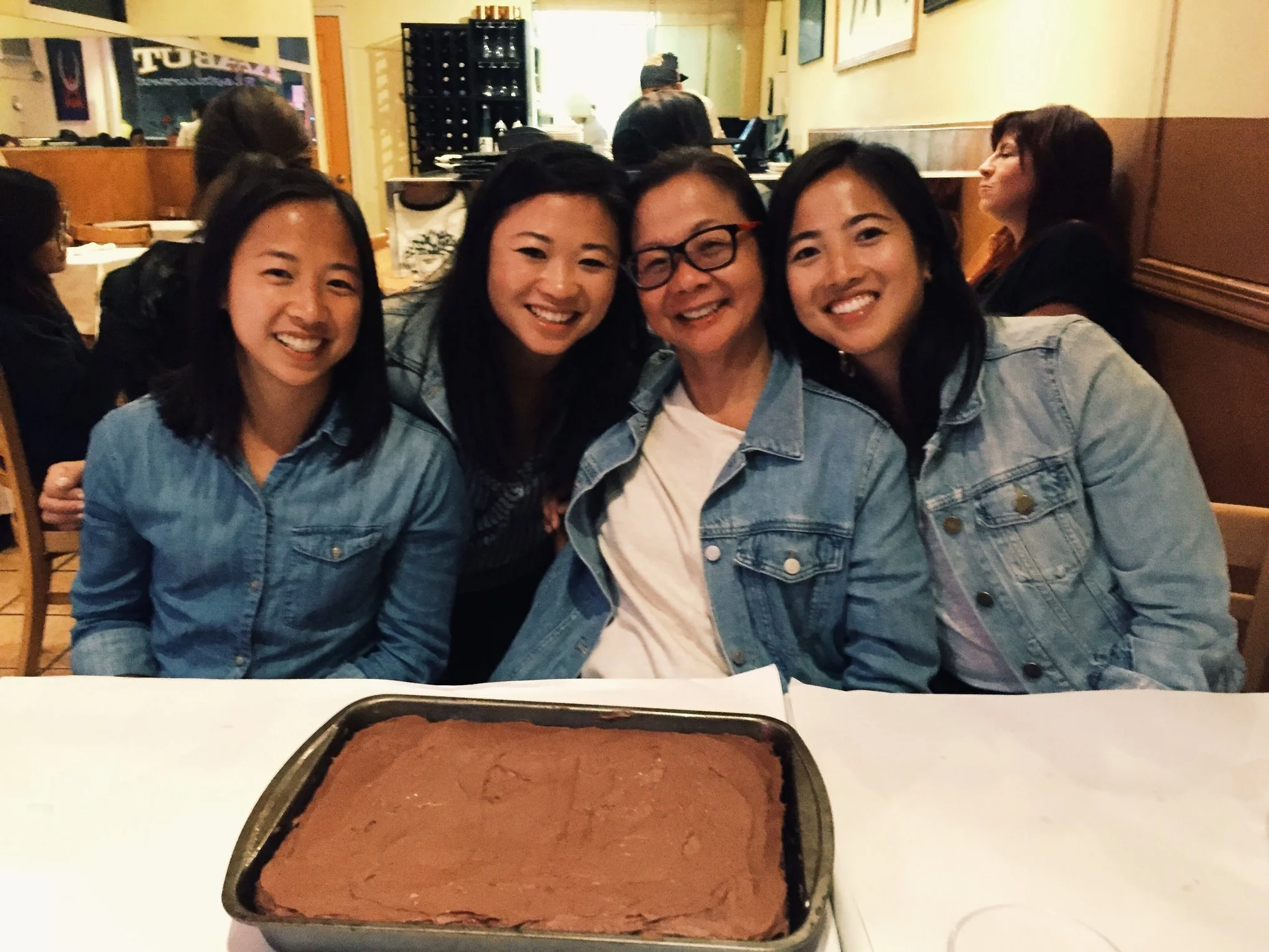 Four women sitting at a restaurant table, smiling and posing for the camera. In the foreground, there is a tray of freshly baked chocolate cake. Background shows other people dining and a wine rack.