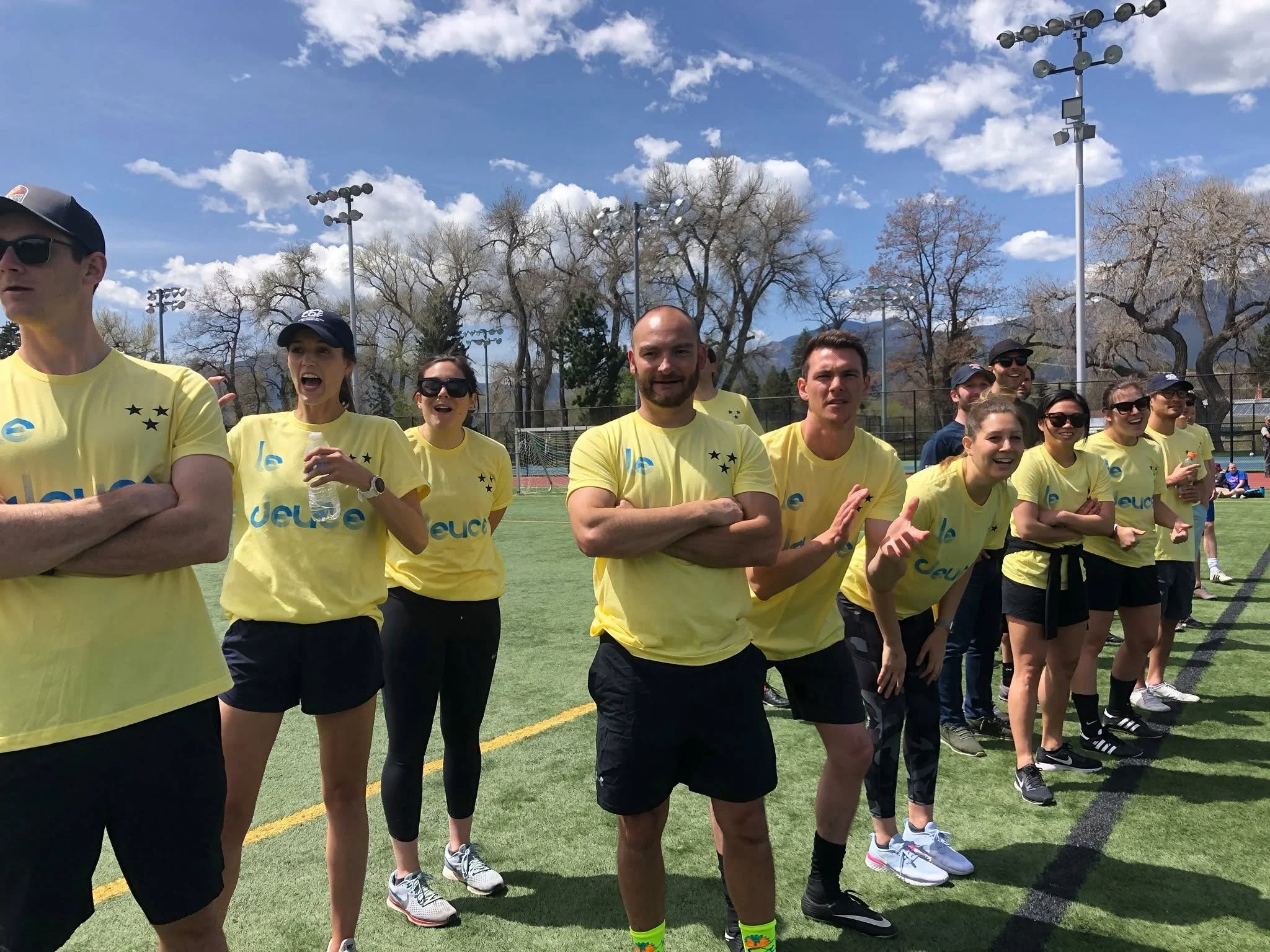 A group of people standing outdoors on a sports field, wearing yellow shirts with a blue logo, with some wearing sunglasses and hats. The sky is blue with scattered clouds, and there are trees and stadium lights in the background.