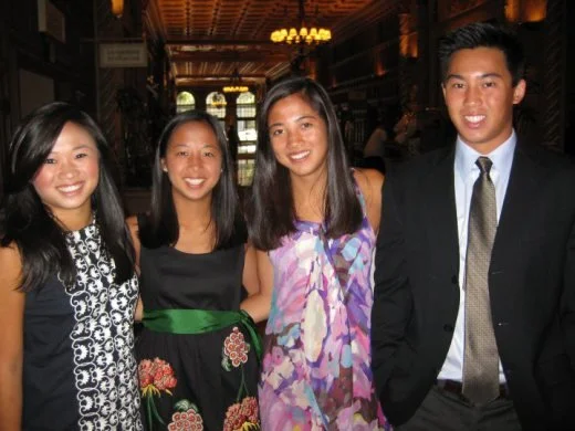 Four young adults, three women and one man, dressed semi-formally, standing together indoors in a warmly lit room.
