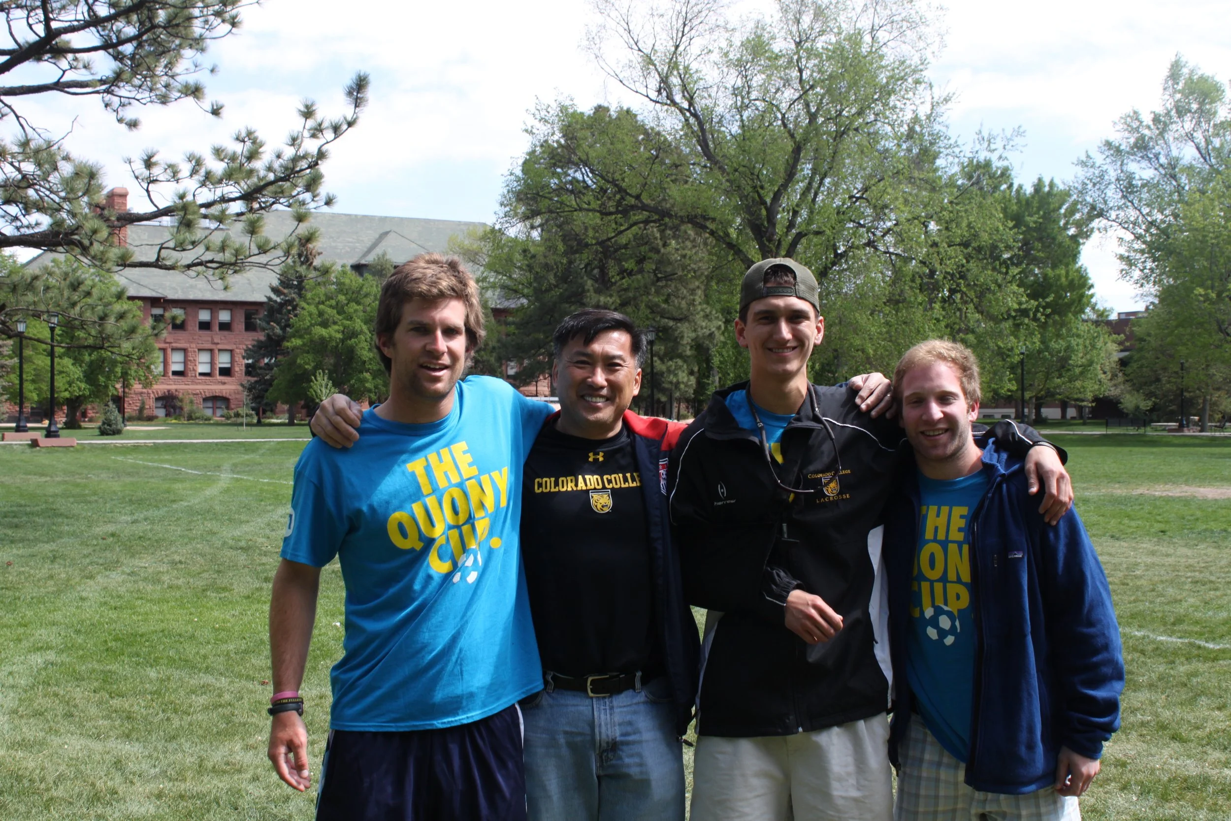 Four young men standing outside on a grassy field with trees and a red brick building in the background, smiling and posing for the photo. They are wearing casual athletic clothing and some shirts say 'The Quarry Cup' and 'Colorado College Lacrosse.'