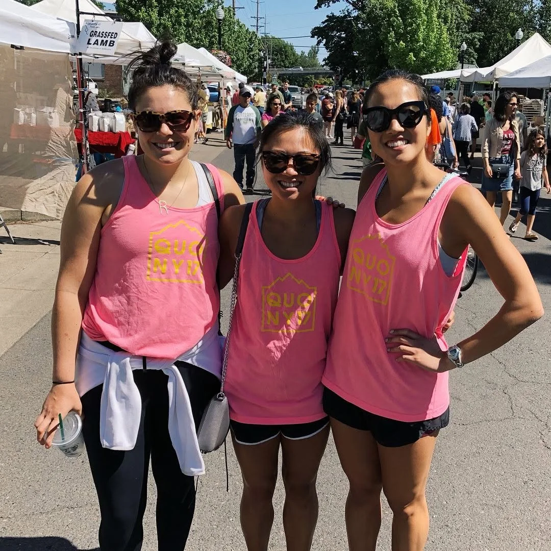 Three women wearing pink tank tops with yellow print, standing outdoors at a street fair or festival, smiling at the camera, with other attendees and vendor booths in the background.