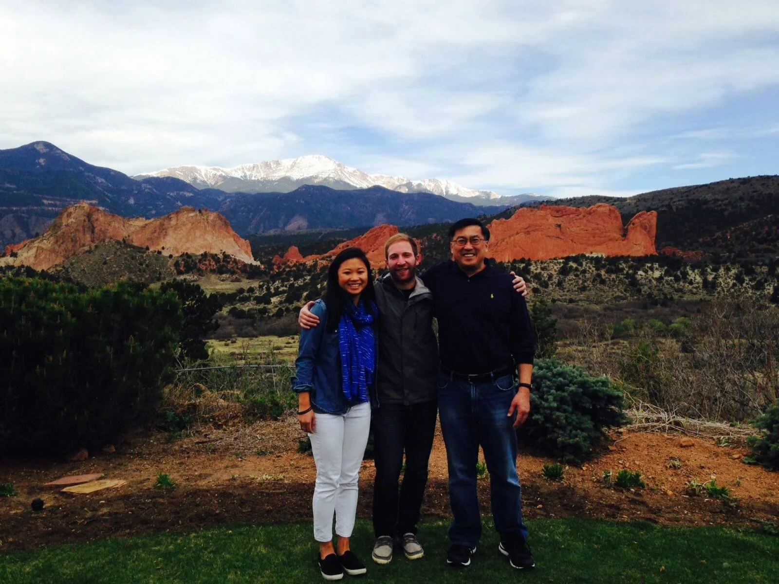 Three people standing together outdoors in front of mountains and red rock formations, smiling, with snow-capped peaks in the background.