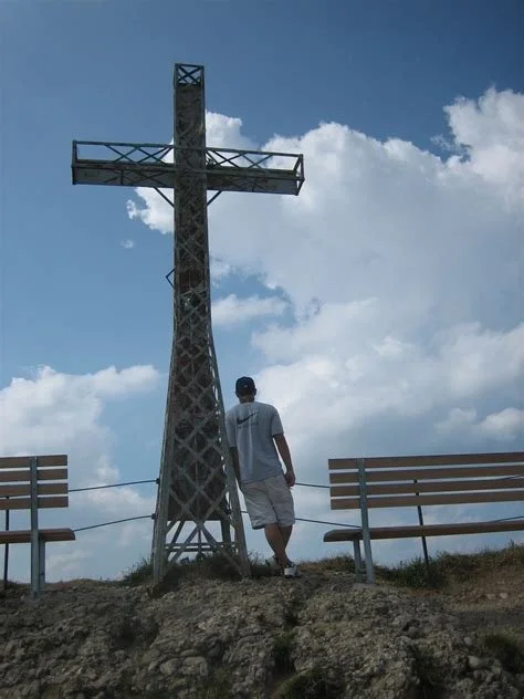 A person standing near a large metal cross monument on a hilltop with two empty benches and a cloudy sky in the background.
