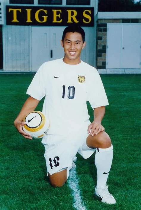 Young male soccer player kneeling on grass field, holding a Nike soccer ball, wearing a white uniform with the number 10, and the team name 'Tigers' in the background.