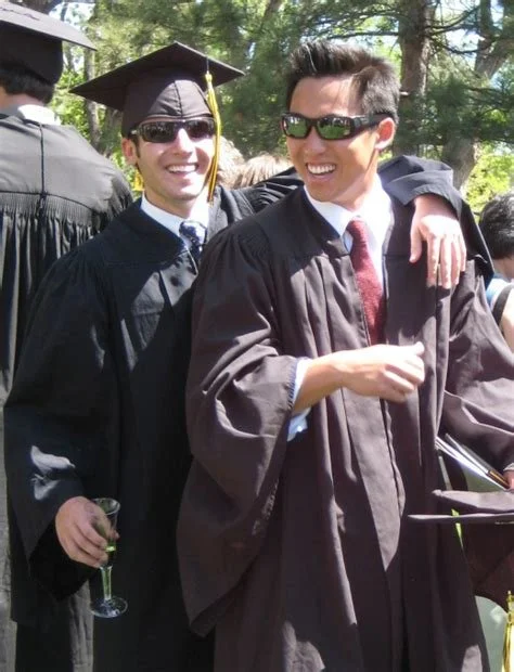 Two young men in graduation caps and gowns smiling, one with a glass of champagne, outdoors under trees.