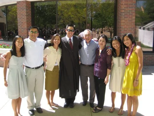 Group of seven diverse people celebrating outdoors in front of a building with glass windows, including a man in a graduation gown and cap.