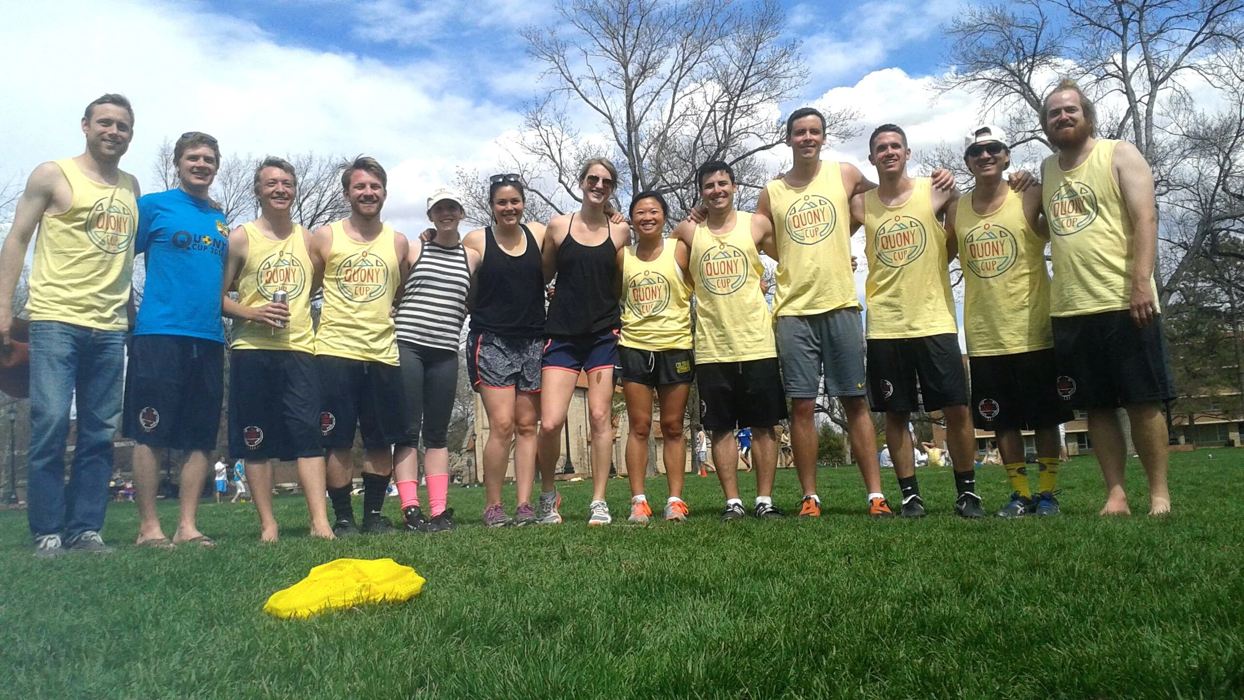A group of people is standing outdoors on grass, smiling with their arms around each other, in a park on a partly cloudy day. Some are wearing yellow tank tops with a logo, and others are in casual athletic or summer clothing.