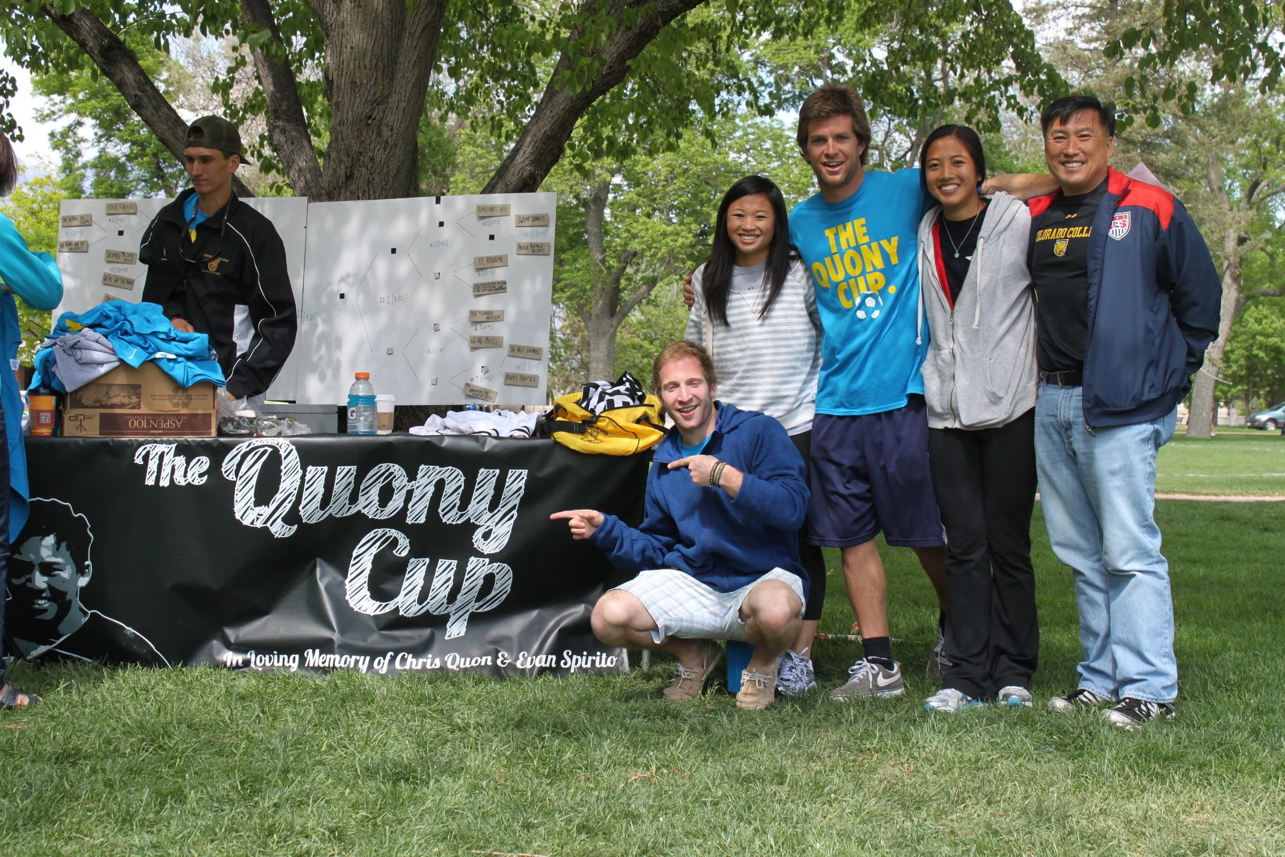 Group of six people smiling and posing together outdoors at a memorial event, with some wearing sports jerseys, under a large tree, with a table and a whiteboard in the background.