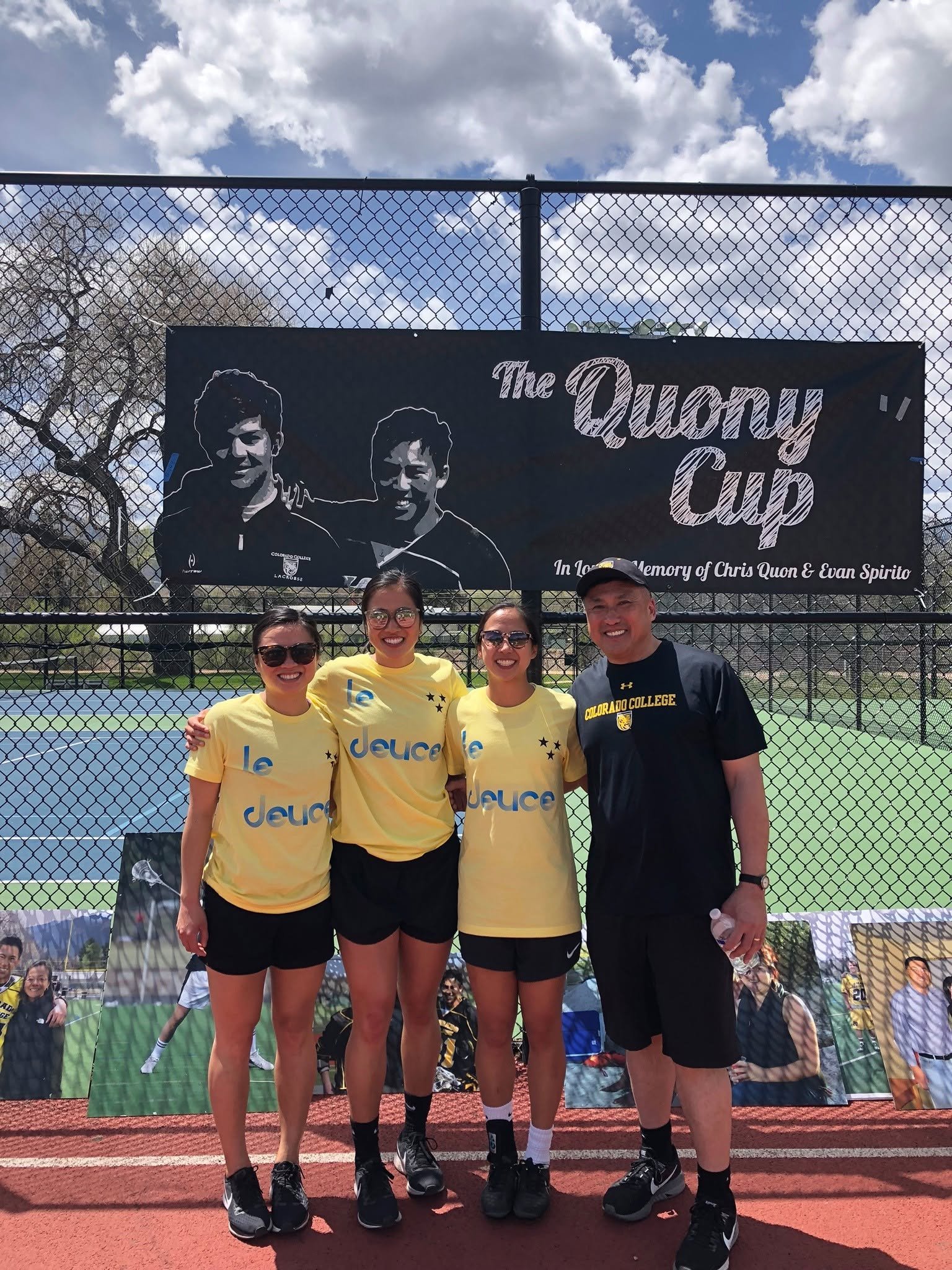 Group of four people posing for a photo at the Quoncy Cup tennis event, standing in front of a black banner with illustrated faces and event name, outdoors on a sunny day with a blue sky and clouds.