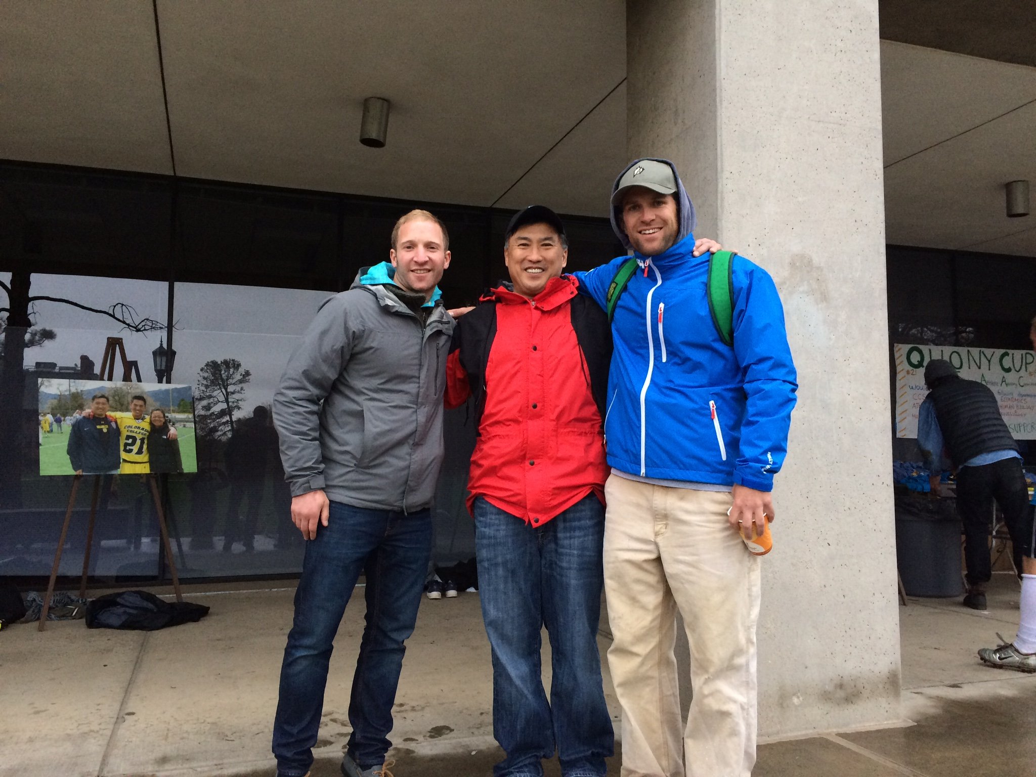 Three men standing together outdoors, smiling, with a building and trees in the background. They are dressed in casual and outdoor clothing, with two wearing jackets and one in a hoodie.