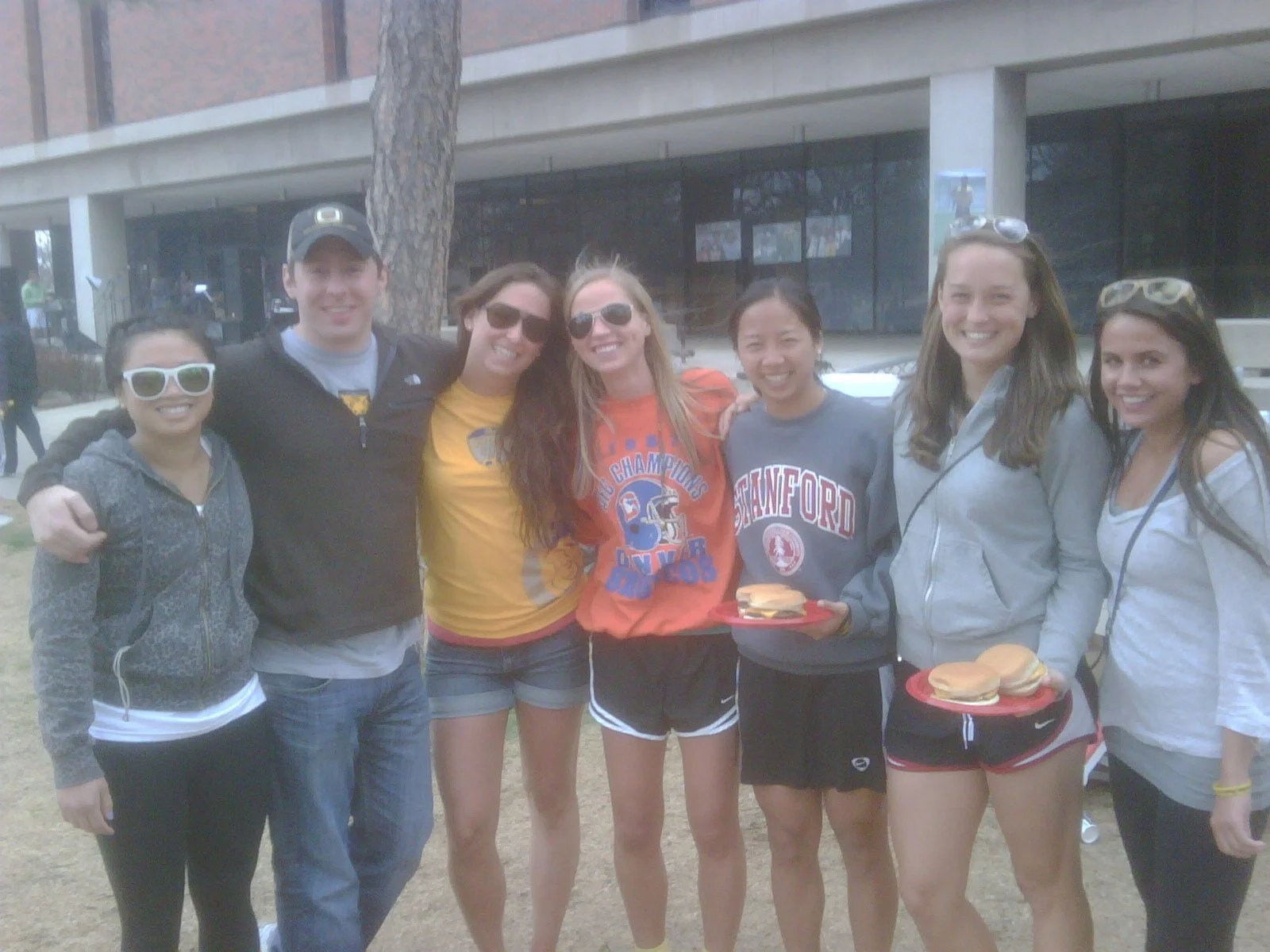 A group of seven young women and one young man standing outdoors, smiling, with some carrying plates of sandwiches, in front of a modern building with large glass windows and a tree.