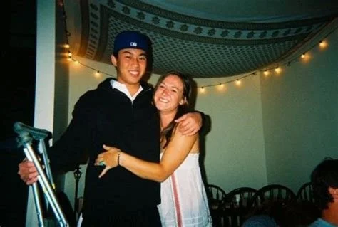 A young man and woman smiling and hugging indoors, with string lights and chairs in the background.