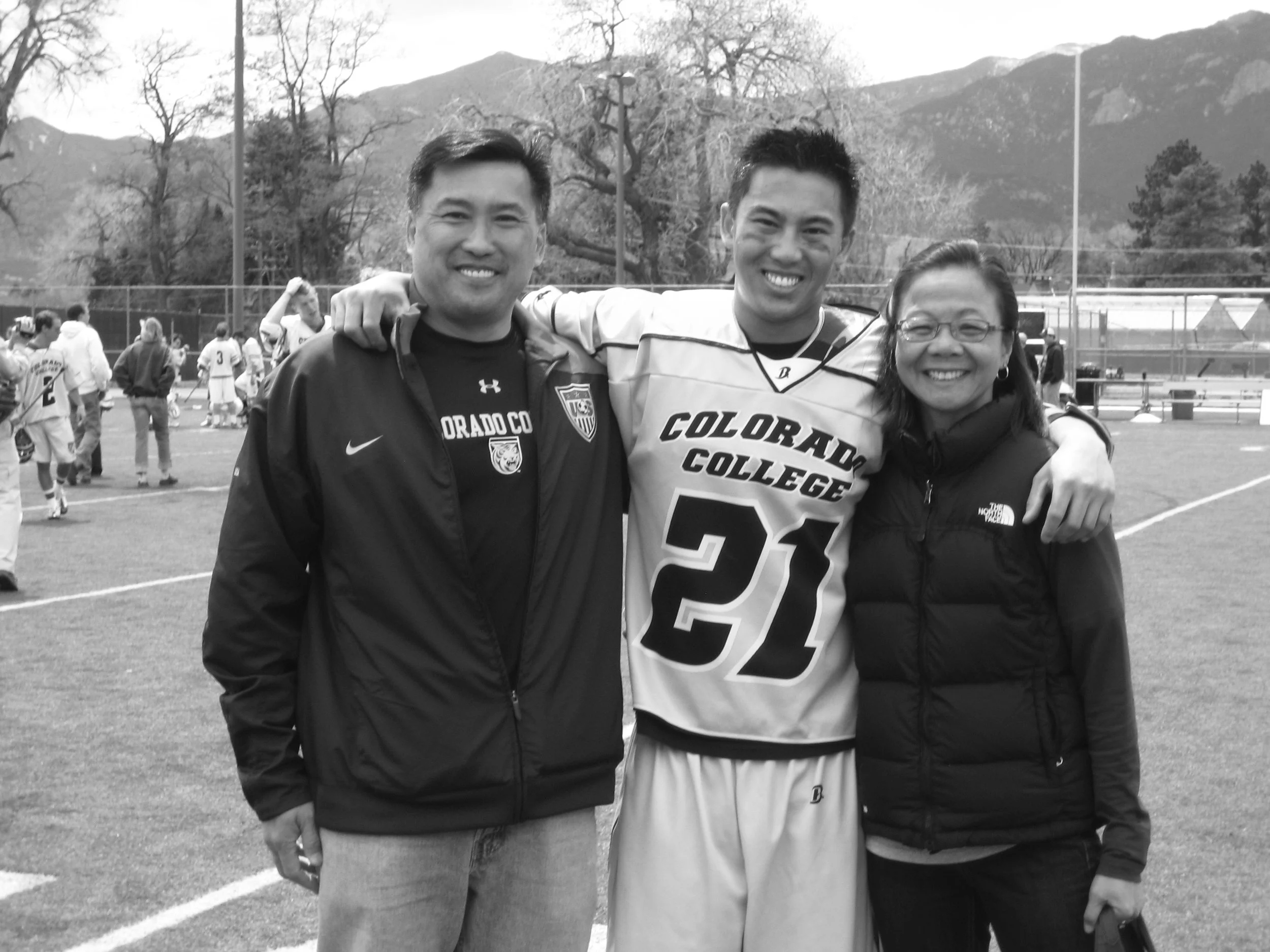 Three people standing on a sports field, smiling, with mountains and trees in the background, black and white photo.