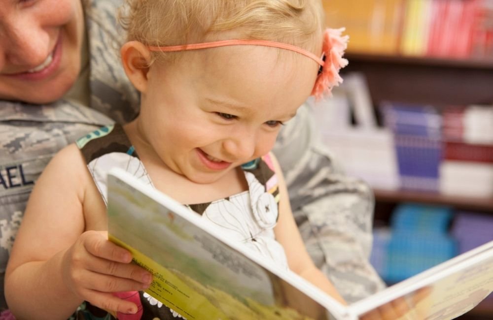 A young girl with a pink headband smiling as she looks at a book, with an adult in a camouflage uniform partially visible behind her.
