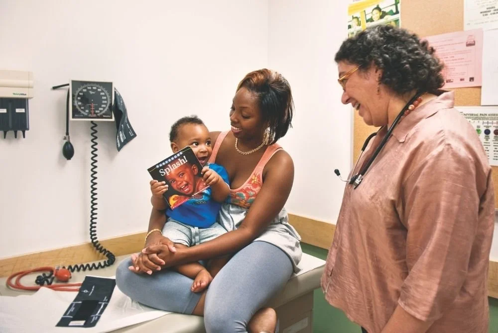 A woman holding a young child sitting on an examination table in a medical office, with a doctor smiling at them. The child is holding a magazine with a picture of a child's excited face. Medical equipment is visible on the wall.
