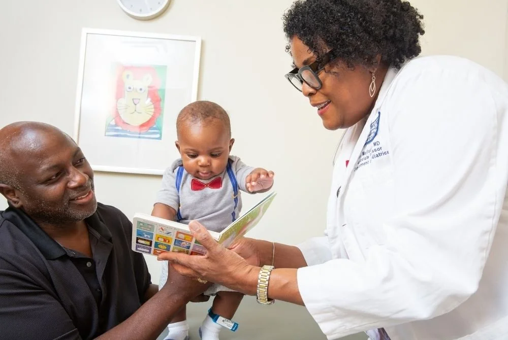 A female doctor showing a children's book to a man and a young child in a doctor's office.