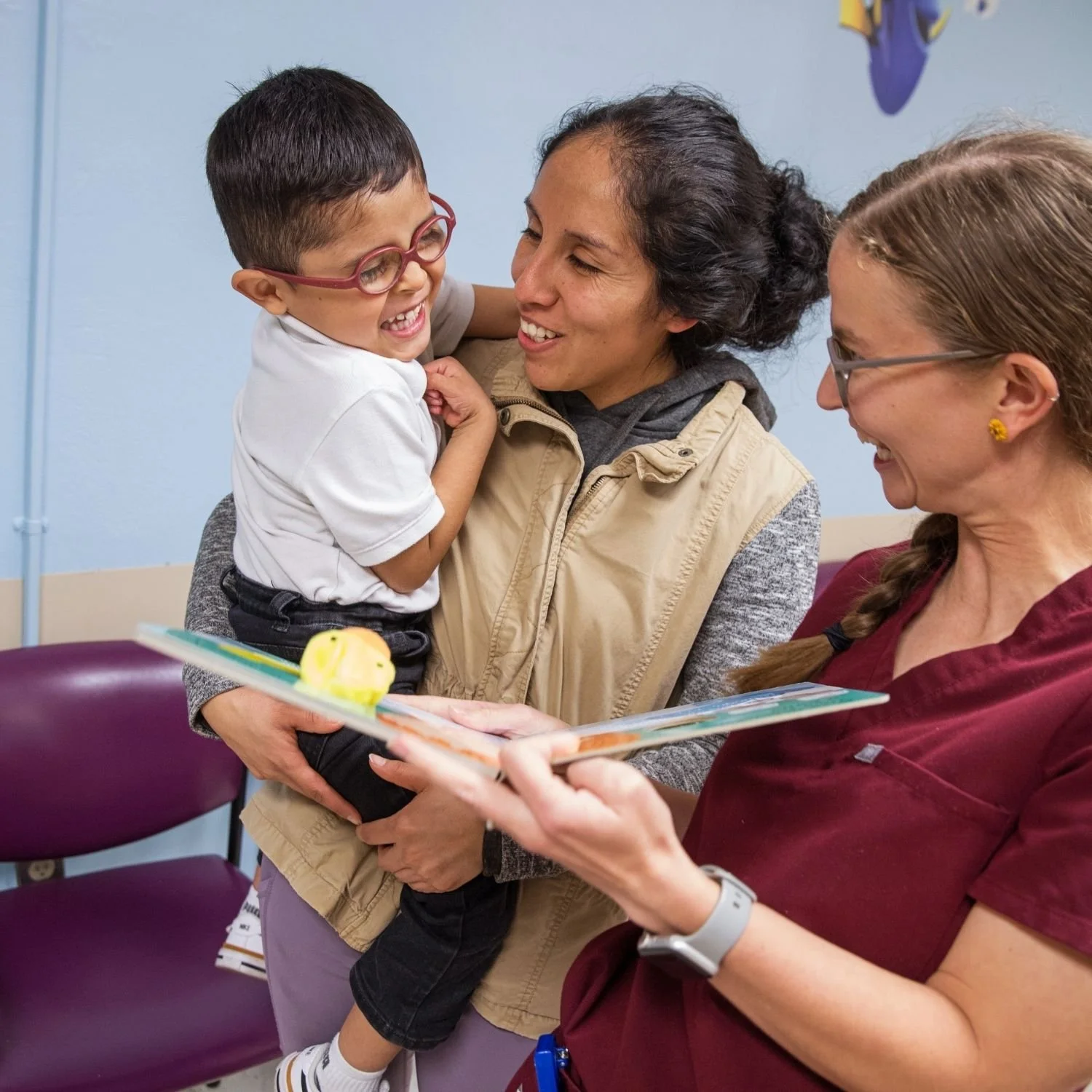 A young boy with glasses being held by a woman, smiling, while another woman in a maroon uniform holds a children's book and looks at the boy, inside a room with blue walls.
