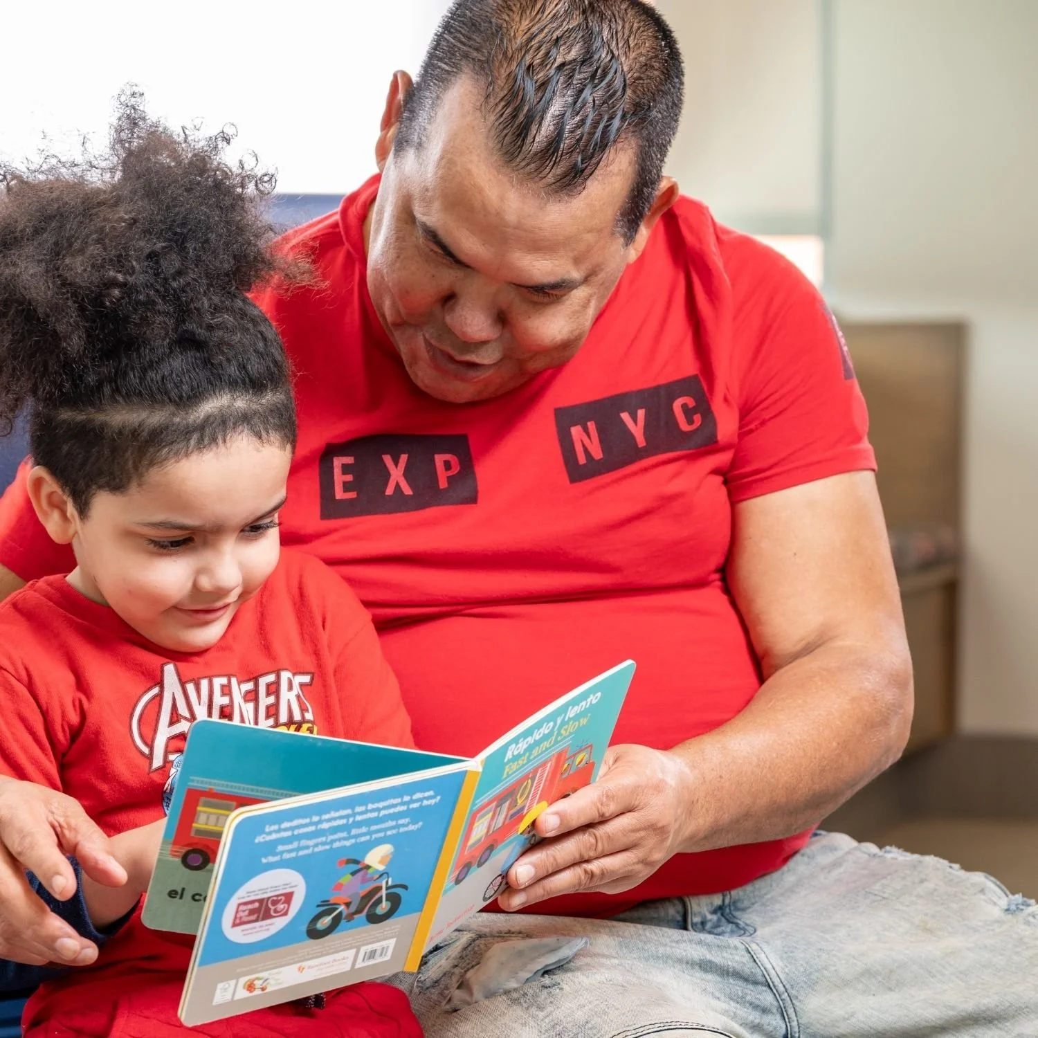 A man and a young girl sitting together, both reading a colorful children's book about transportation, with the man wearing a red T-shirt and the girl wearing a red Avengers T-shirt.