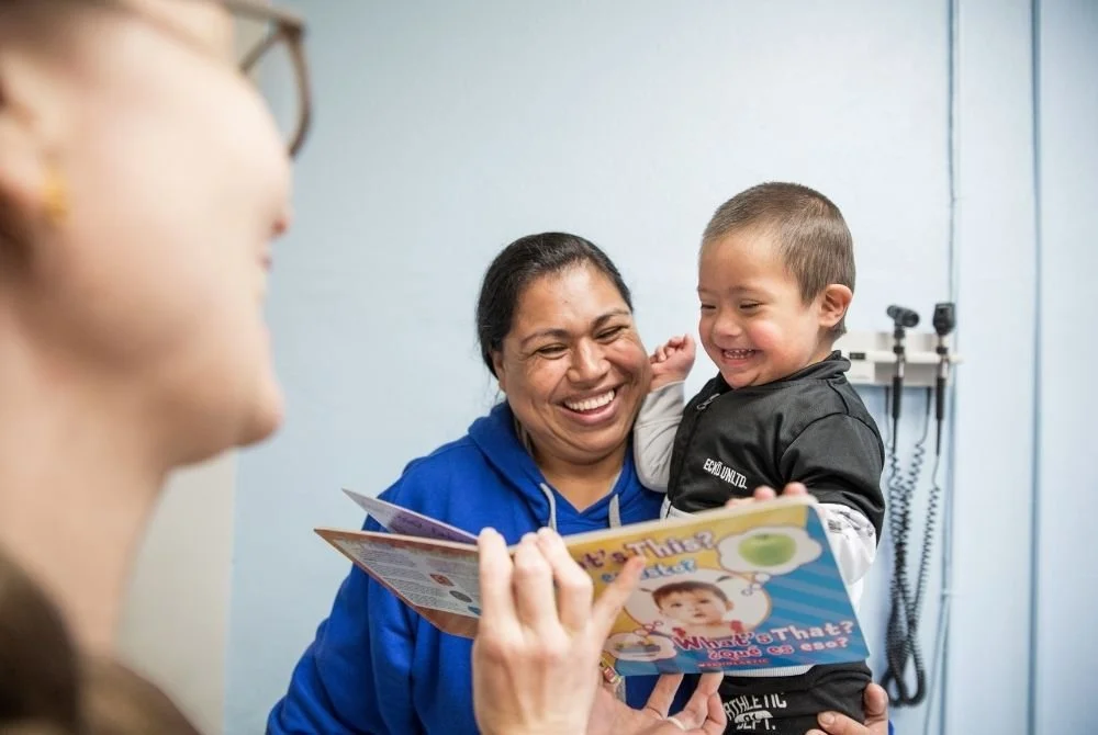 A woman in a blue hoodie holding a young boy in a black jacket, both smiling and looking happy during a visit to a healthcare provider's office. A blurred face of a healthcare professional holding a pamphlet is visible in the foreground.