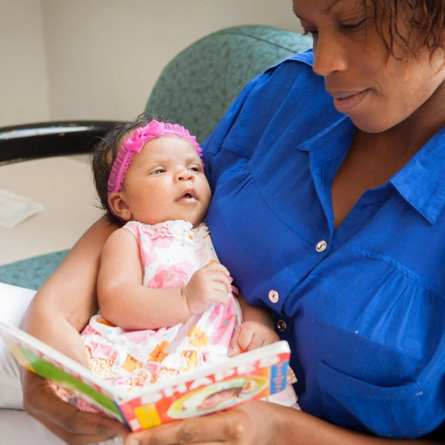 A woman holding a baby girl in her arms during a visit to the hospital, with the woman wearing a blue shirt and the baby dressed in a pink and white floral outfit with a matching pink headband.