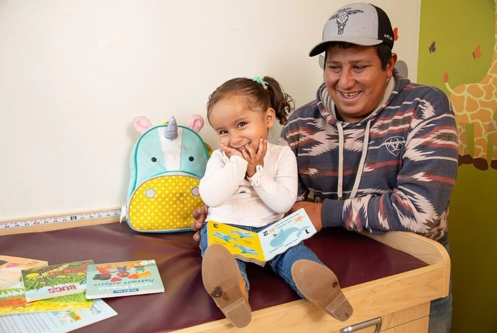Young girl and man smiling while sitting on a table with children's books. Girl holds a shark shape toy, girl has a unicorn bag in the background.