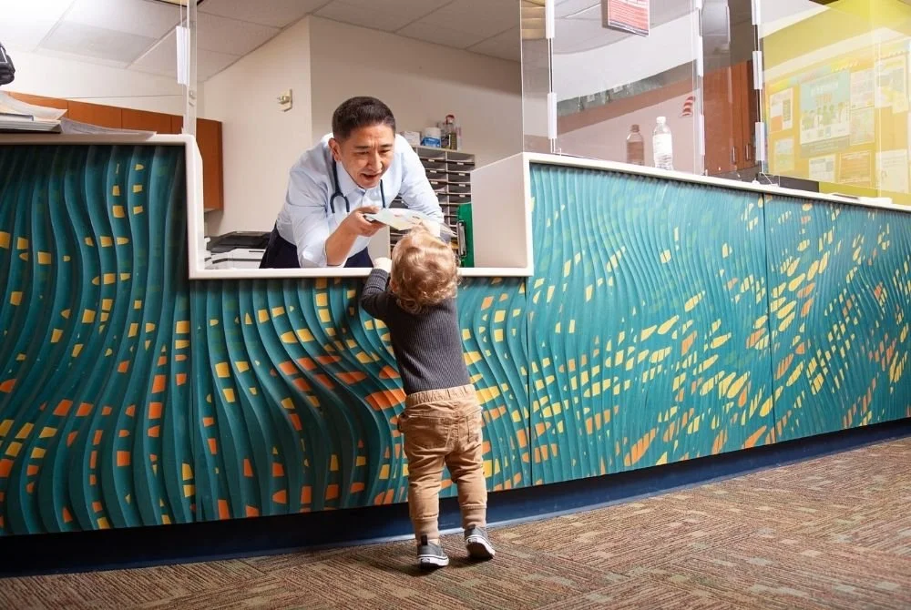 A young child with curly hair, wearing a dark sweater and brown pants, standing at a hospital or clinic reception desk, handing a paper to a healthcare worker behind the counter. The counter has a colorful, wavy design.