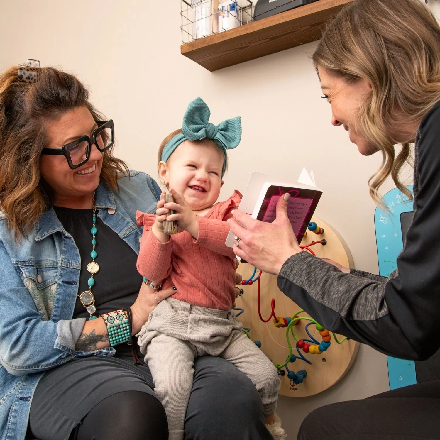 A young girl with a big blue bow headband smiling and laughing during a medical exam, held by a woman with glasses and tattoos, while another woman showing her a card.