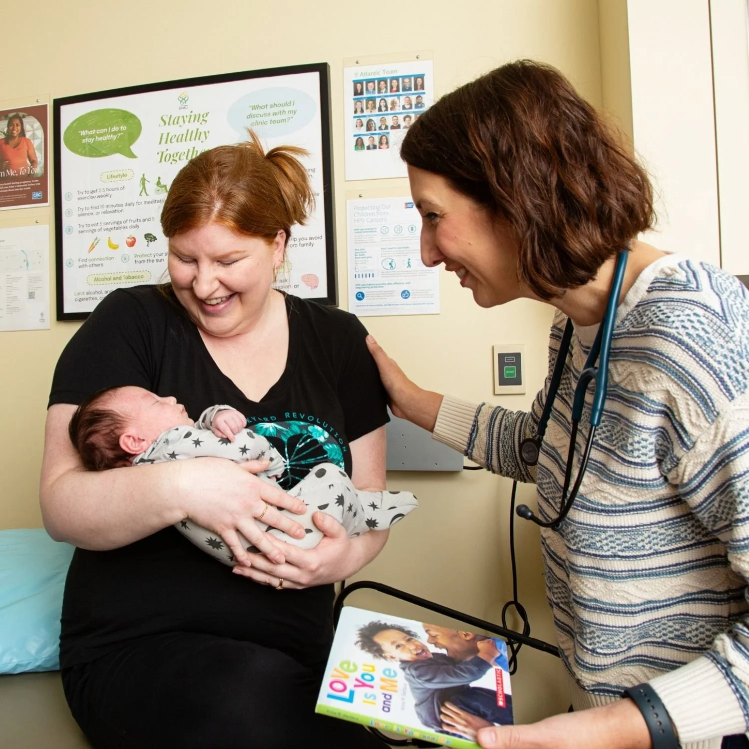 A woman holding a newborn baby in a medical office. A healthcare professional with a stethoscope on her neck is smiling and talking to the woman. Posters and informational materials are on the wall.