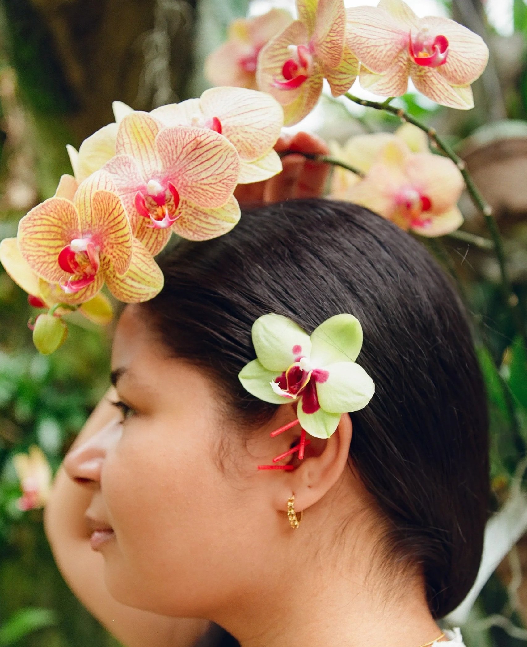 A woman with acupuncture needles and dark hair wearing orchids behind her ear, jewelry, and traditional attire, seen in profile against a blurred greenery background.