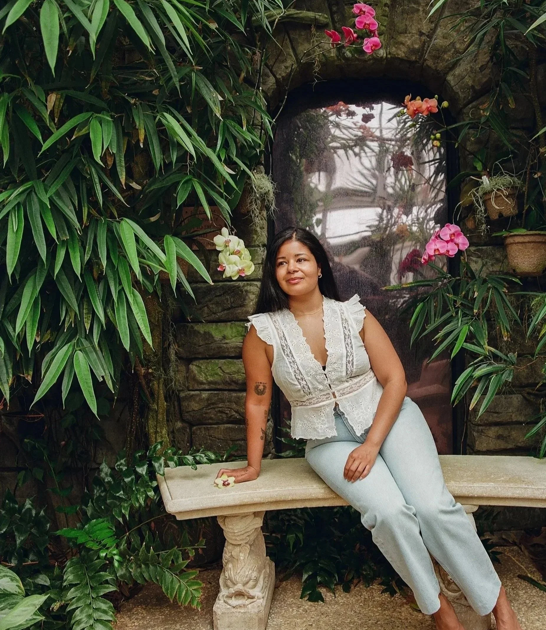 Woman sitting on stone bench in lush garden filled with orchids and green foliage, with stone arch and vines in the background.