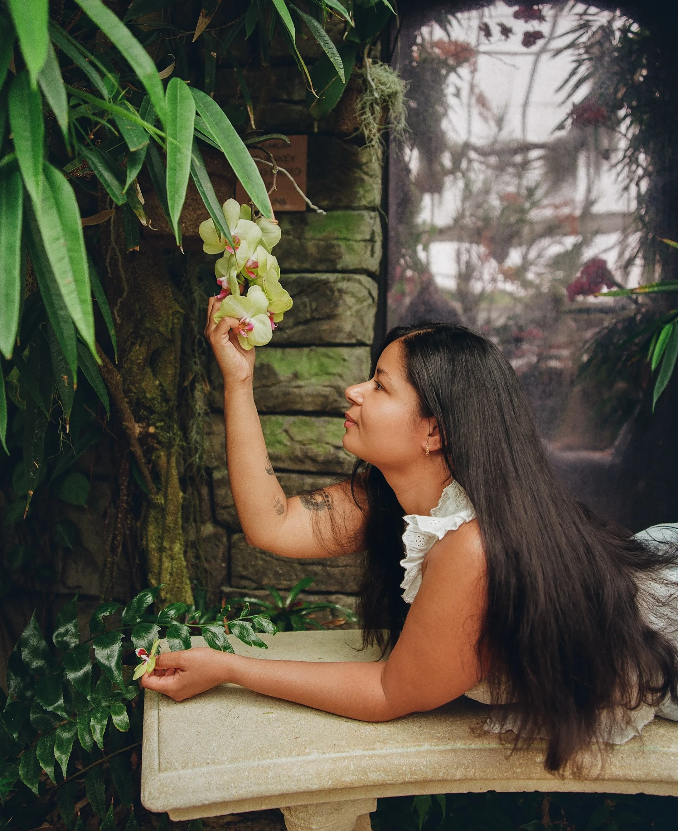 A woman with long dark hair and tattoos on her arm, dressed in white, is reaching up to a light yellow orchid flower on a plant on a stone wall in a garden.