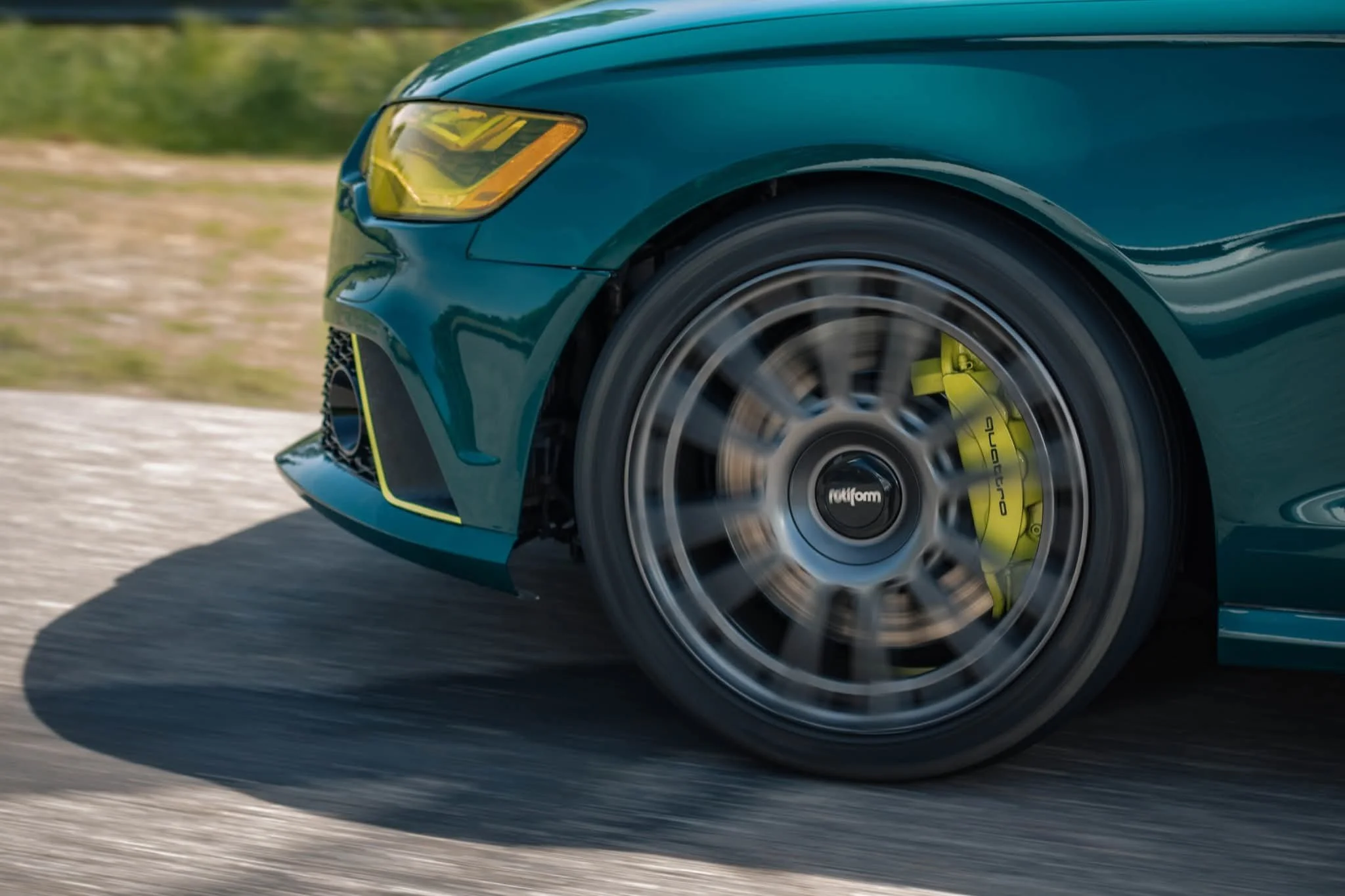 Close-up of a teal sports car's front end and wheel in motion on a road, with blurred background.