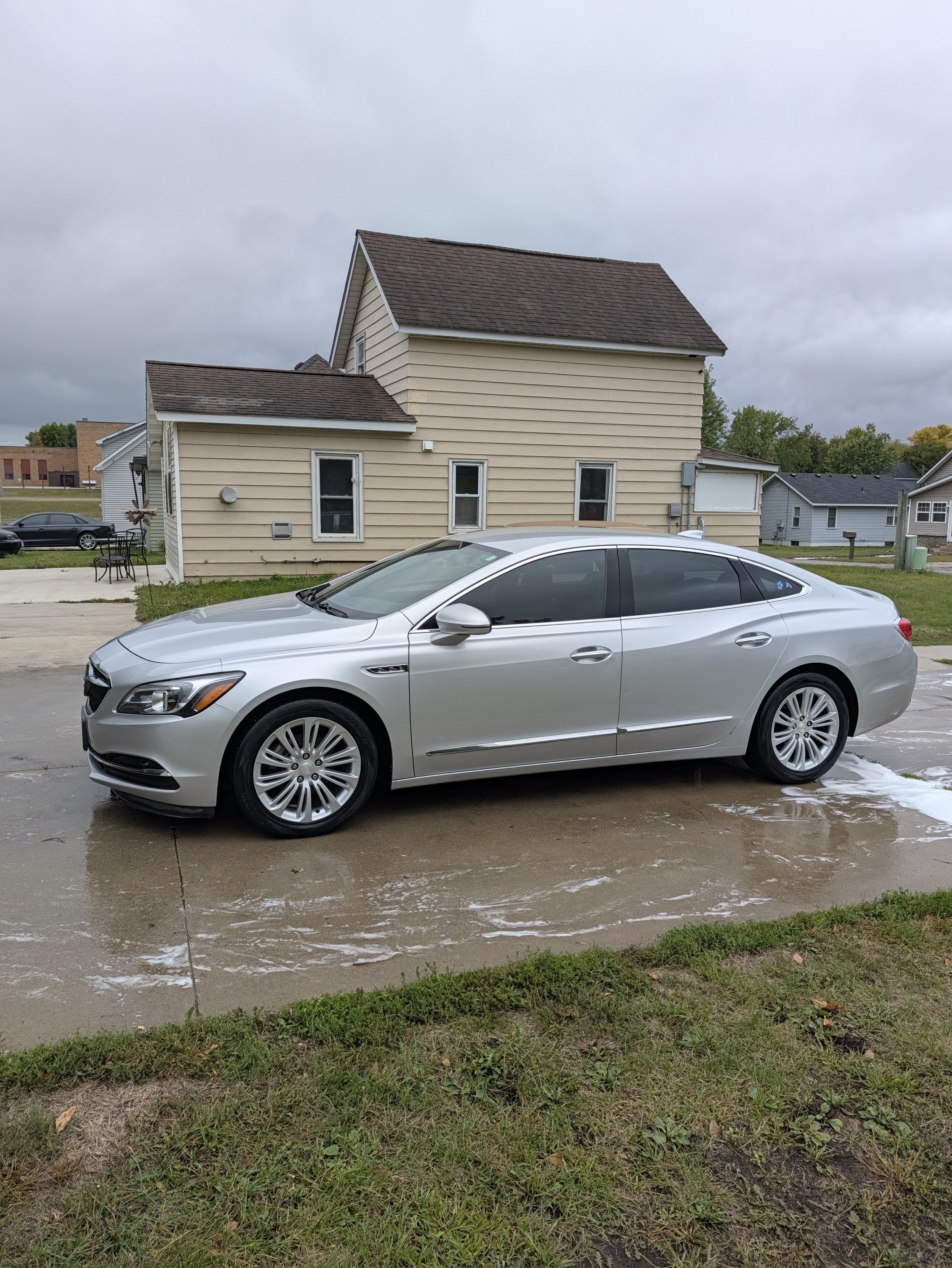 A silver car parked on a wet driveway in front of a beige house with a small lawn and neighboring houses, under a cloudy sky.