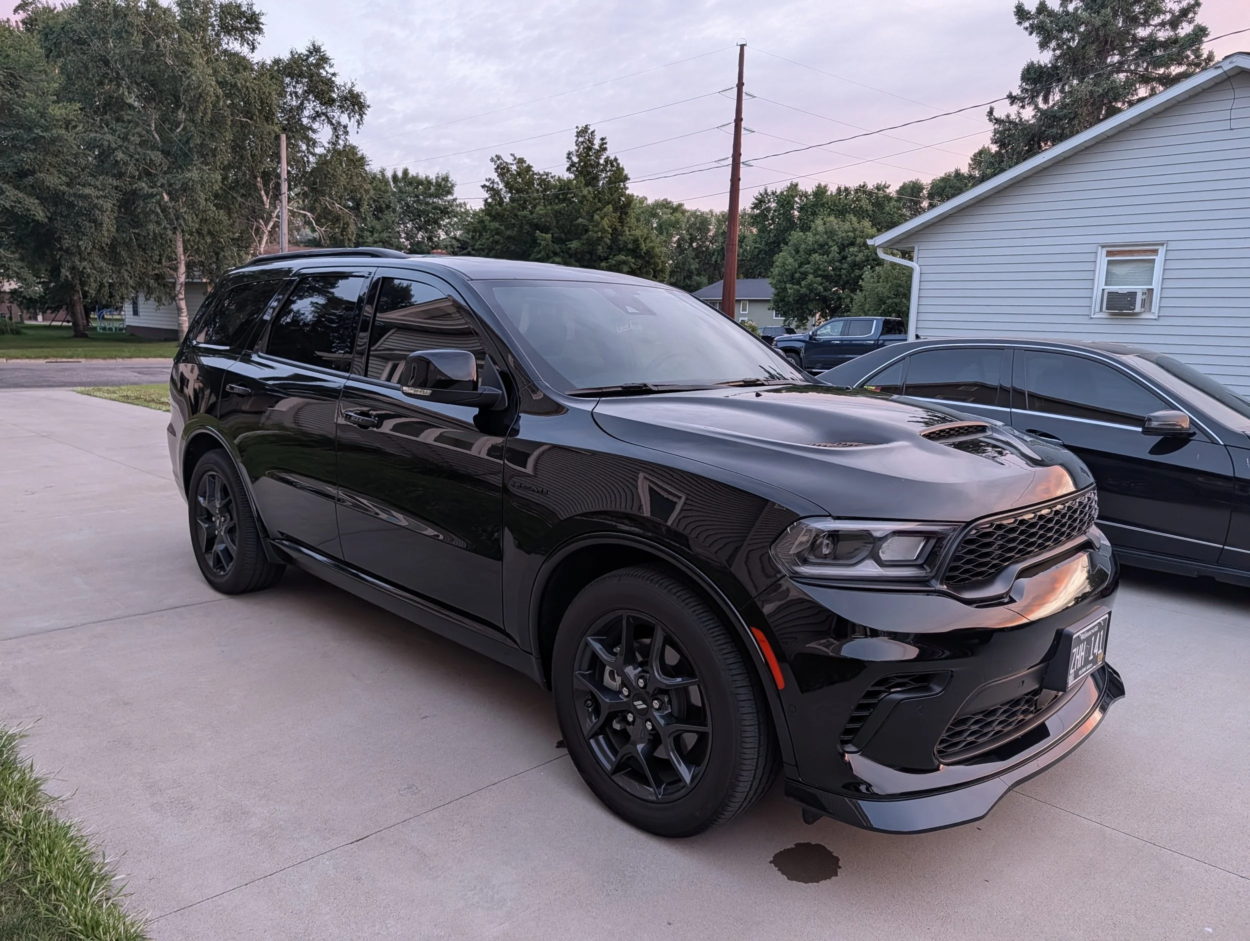 A black SUV parked on a concrete driveway with other cars and a house in the background, surrounded by trees and utility poles, during dusk.