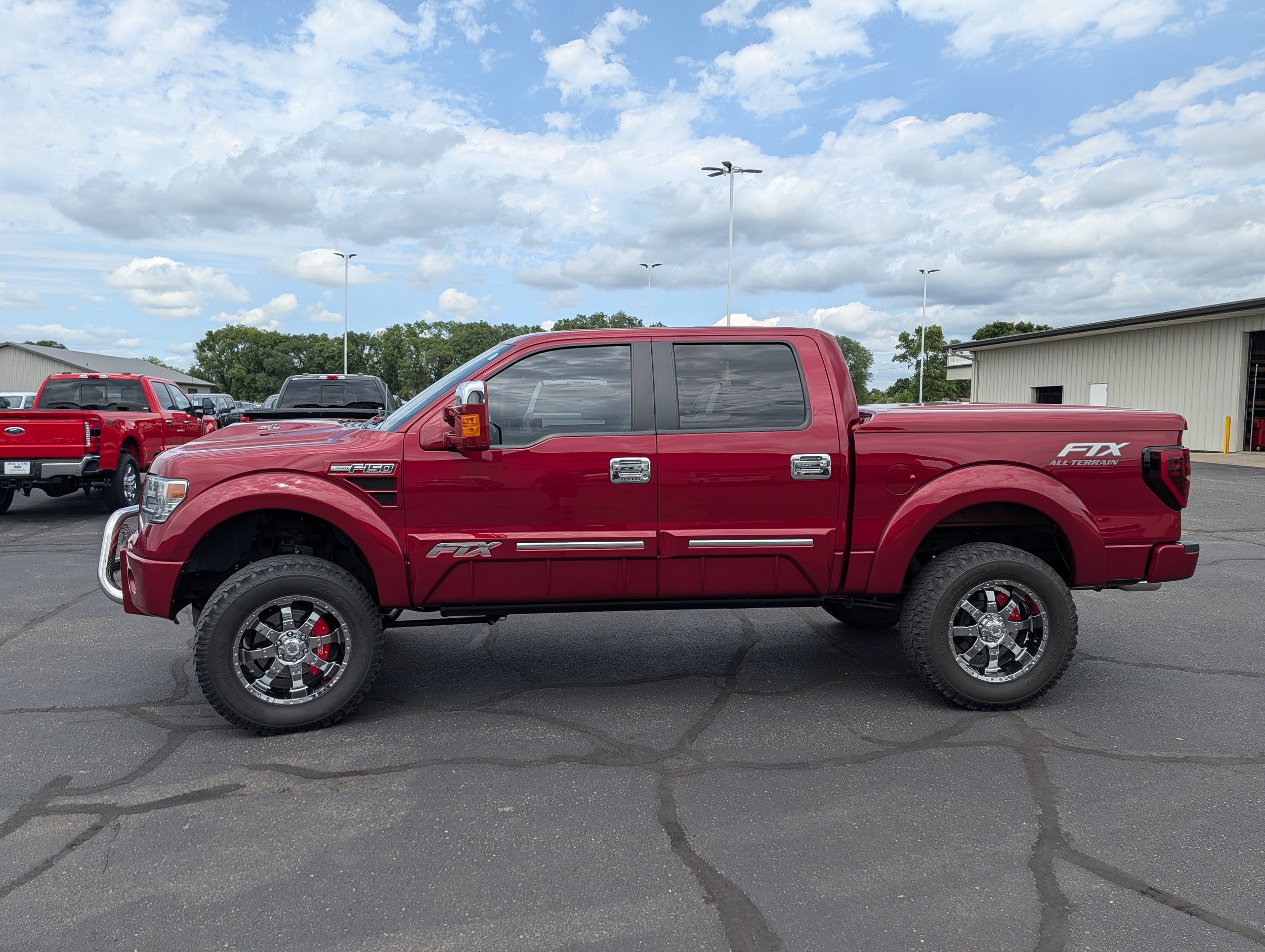 A red Ford F-150 all-terrain pickup truck parked in a lot with other vehicles, under a partly cloudy sky.