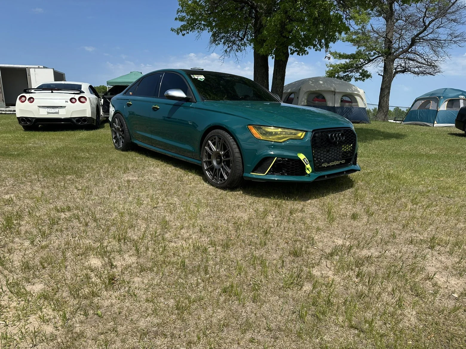 A teal Audi sedan parked on grass at an outdoor car show. In the background, there is a white sports car, tents, and trees under a blue sky.
