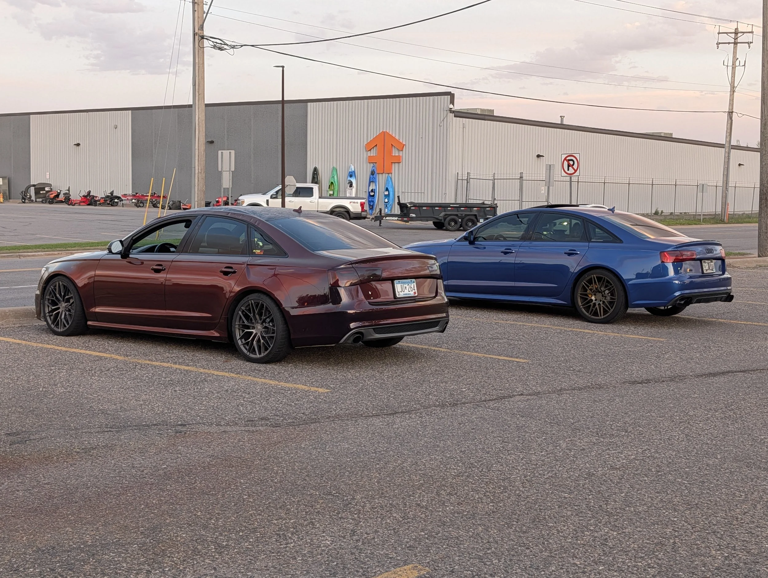 A parking lot with two cars in the foreground, a building with a large orange symbol, kayaks on the side of the building, and power lines overhead during sunset.