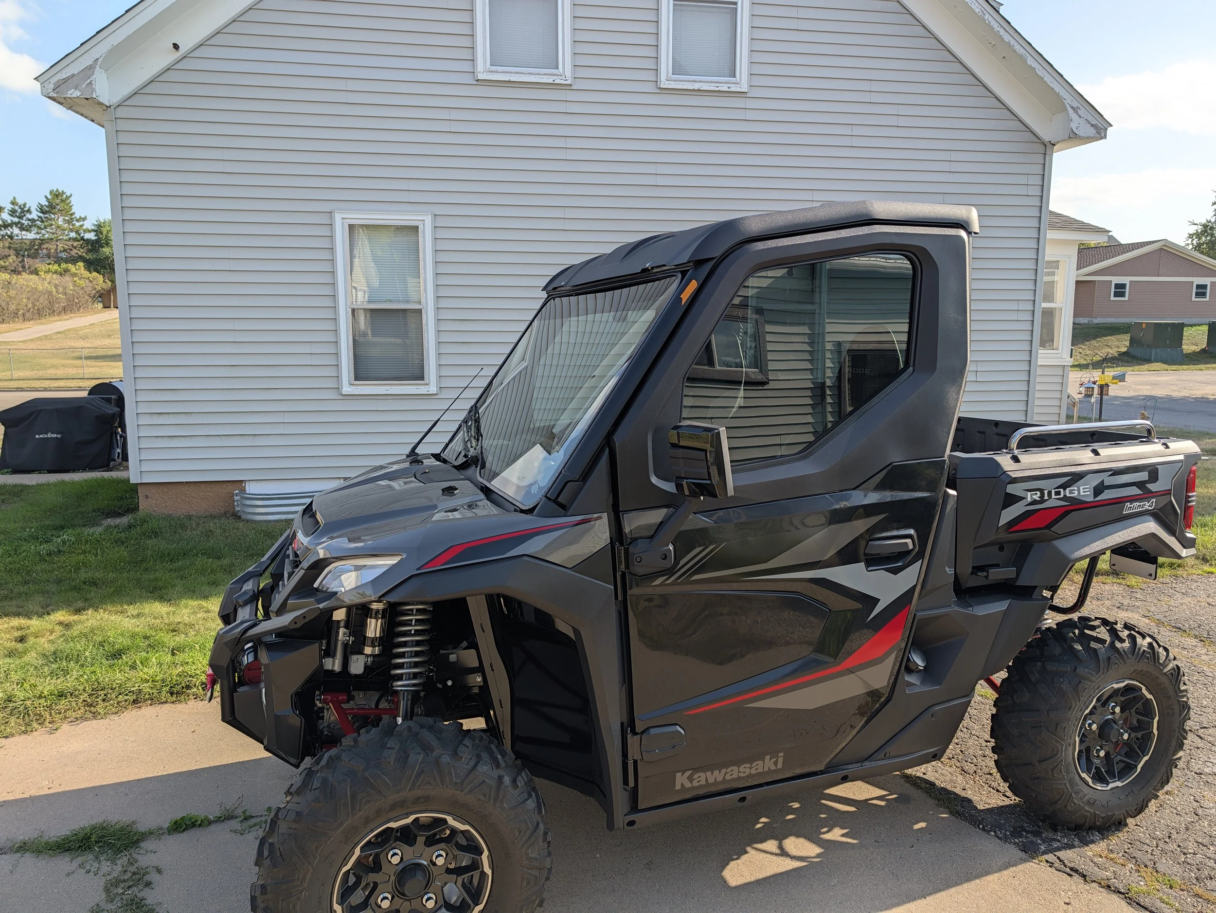 A black Kawasaki off-road utility vehicle parked on a sidewalk in front of a white house with beige siding, several windows, and a well-maintained lawn.