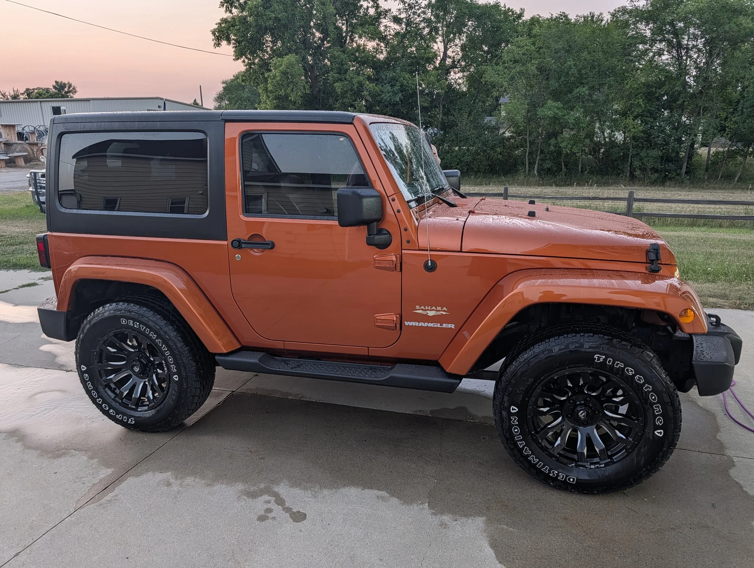 Orange Jeep Wrangler Sahara parked on a concrete driveway with water droplets on its surface, surrounded by lush green trees, and a metal building in the background at dusk.
