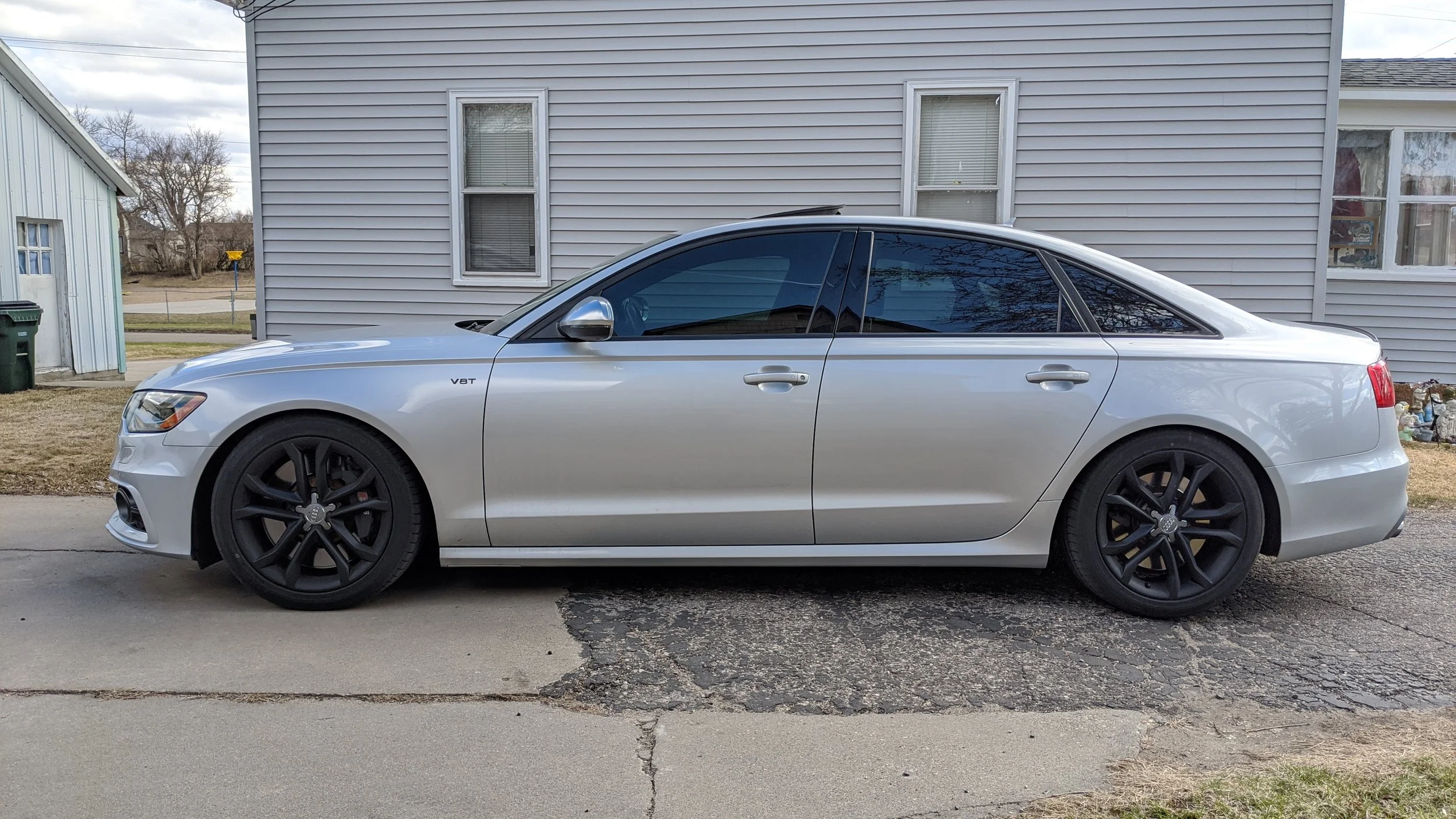 Silver coupe car parked in front of a light gray house with vinyl siding and two windows with blinds.