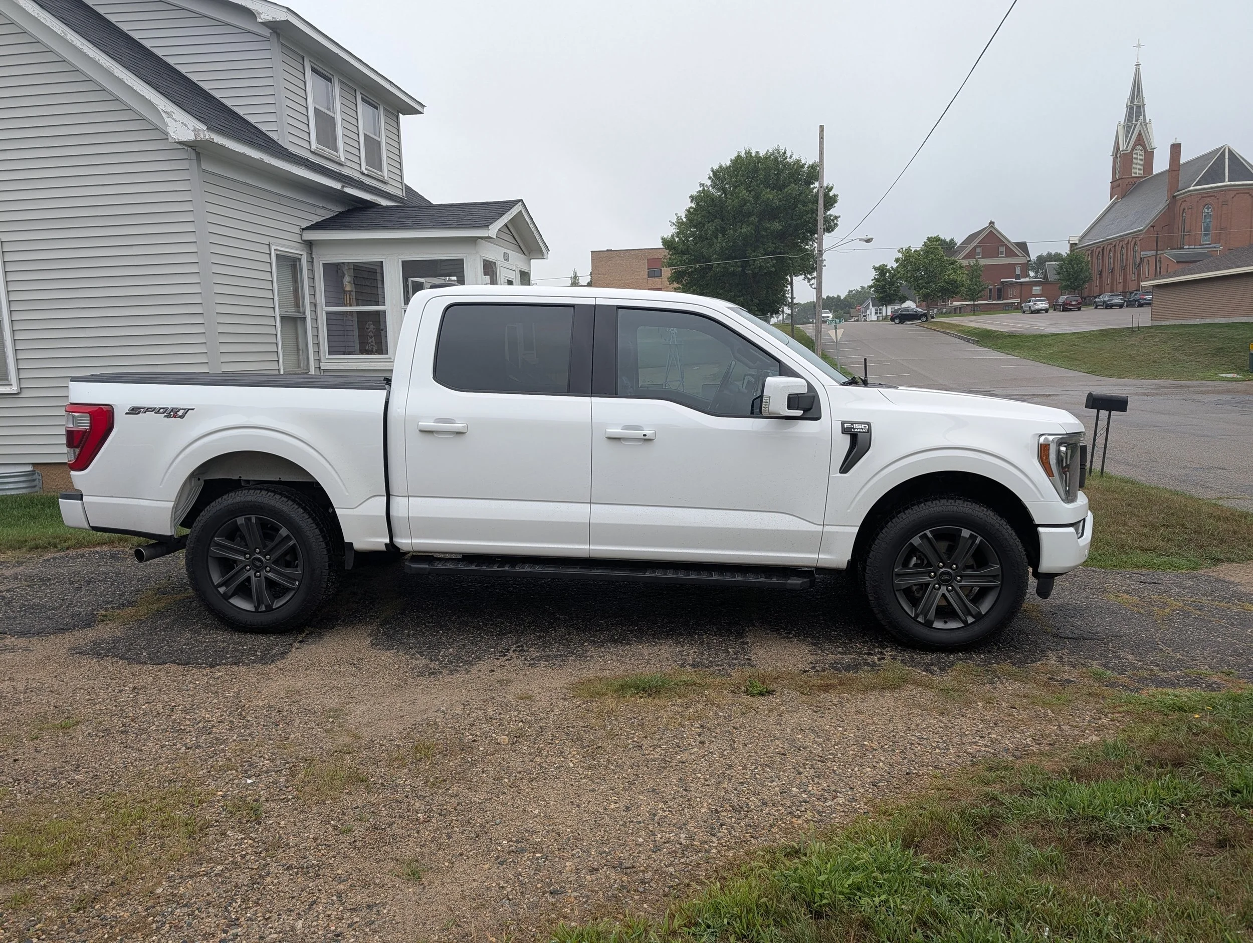 A white pickup truck parked on a gravel surface in front of a beige house with vinyl siding, with a church and other buildings in the background on a cloudy day.