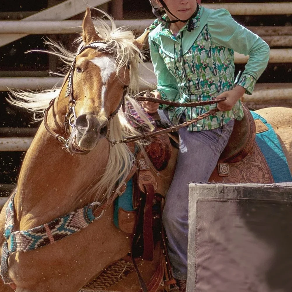 A young girl riding a brown horse with a blonde mane, wearing a green jacket, blue jeans, and a helmet, at a riding arena.