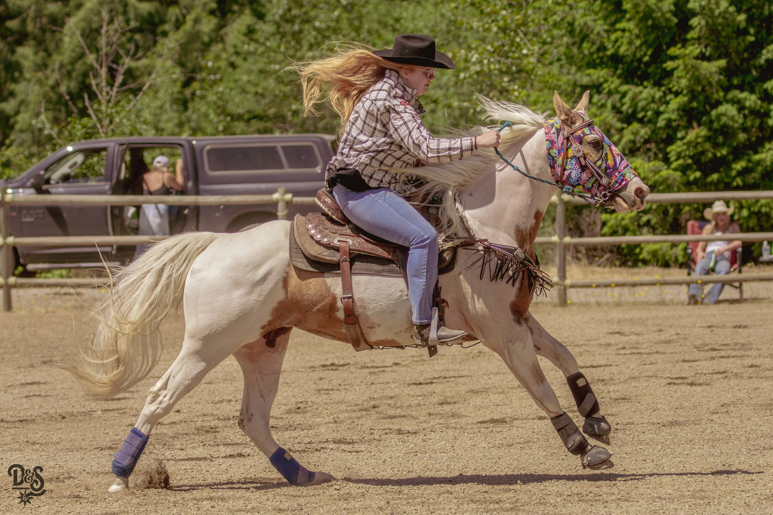 Woman riding a white horse with a colorful saddle and bridle, wearing a black hat, plaid shirt, and jeans, at an outdoor equestrian event.