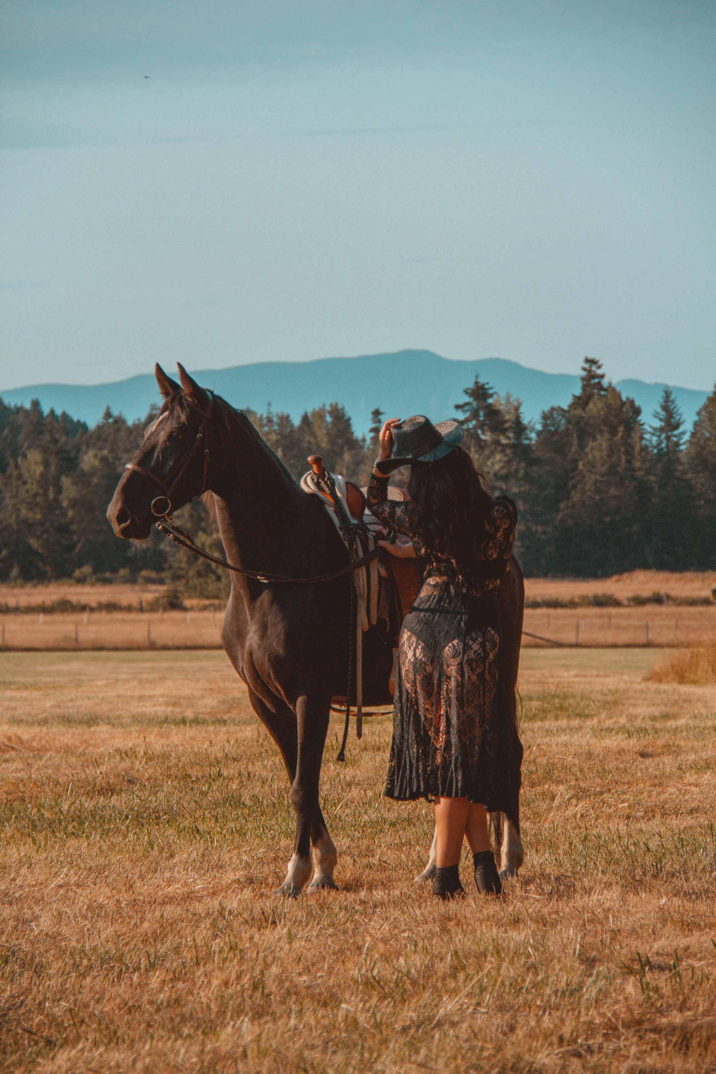 A woman in a black lace dress with a hat standing next to a brown horse in an open field with mountains in the background.