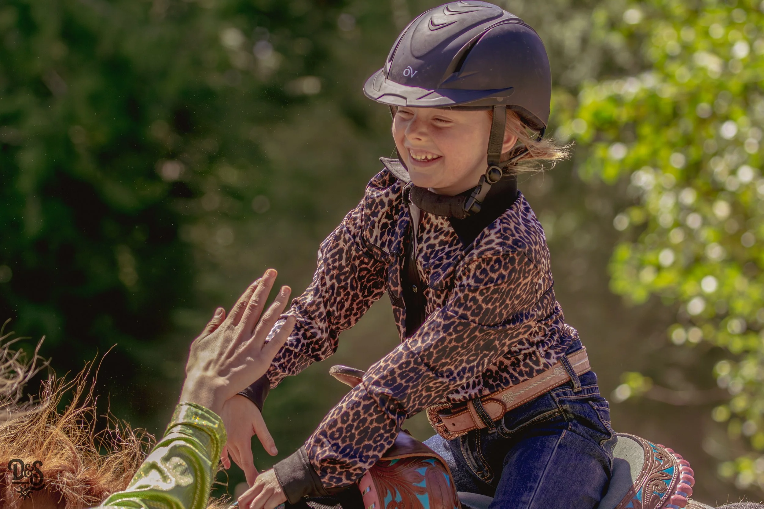 A young girl riding a horse and smiling, wearing a helmet and animal print jacket, outdoors with green trees in the background, reaching out to high-five someone.