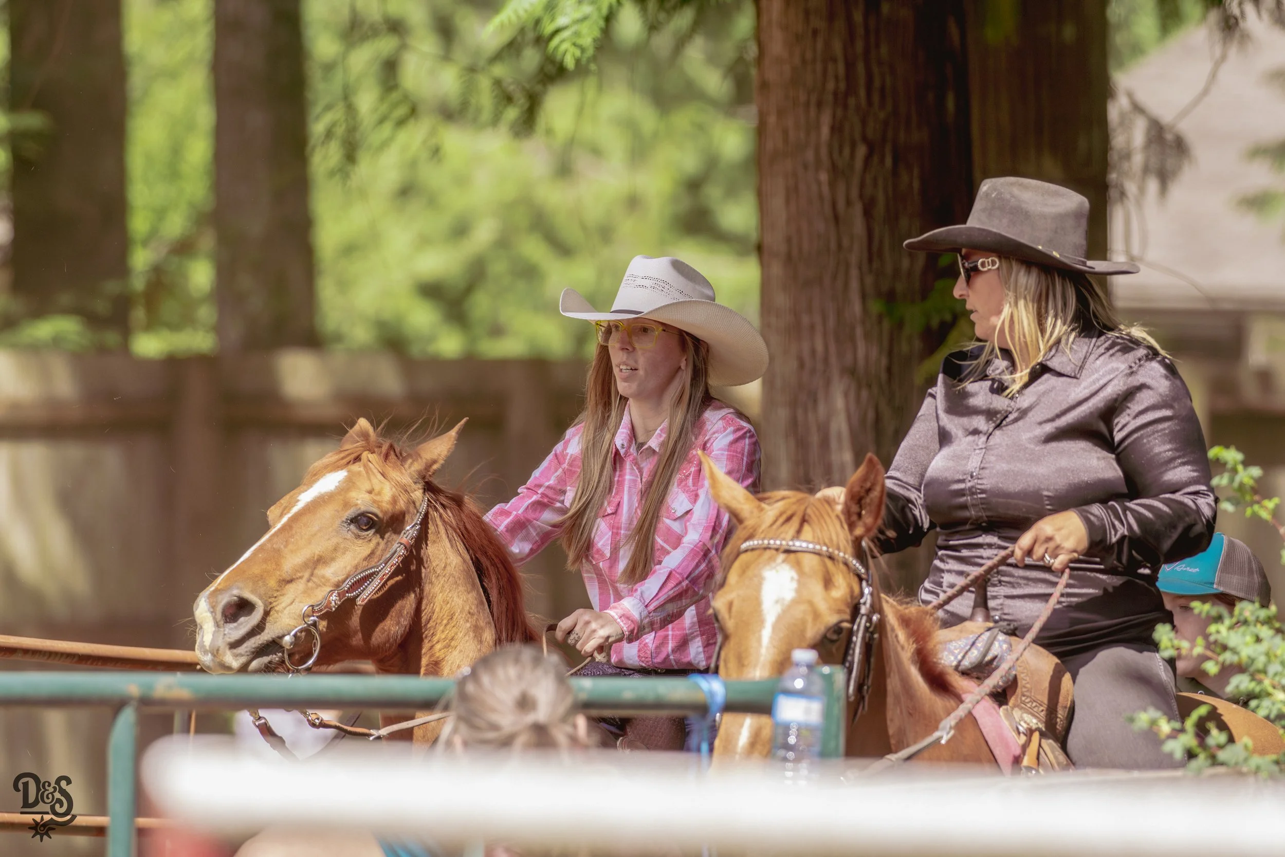Two women riding horses outdoors among tall trees, wearing cowboy hats and sunglasses, with other riders partly visible in the background.
