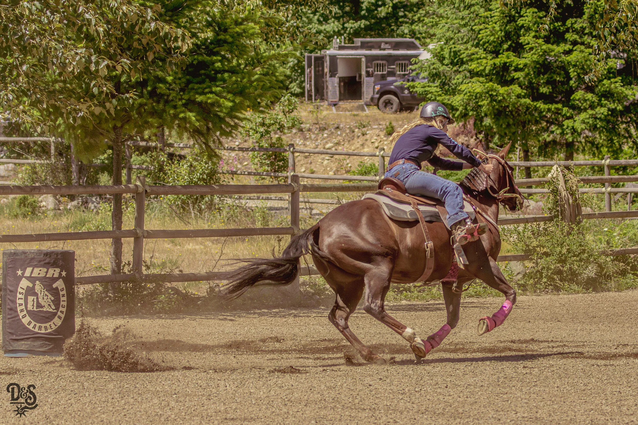 A person riding a horse on a dirt track in an outdoor riding arena surrounded by trees and greenery.